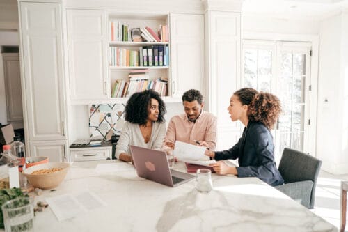 The image shows a professional real estate agent, likely discussing documents with a couple at a kitchen table. The agent appears to be explaining details, while the couple listens attentively. The setting is casual yet professional, emphasizing an atmosphere of trust and open communication. This could illustrate a moment where the agent is building trust with clients through transparency and guidance.