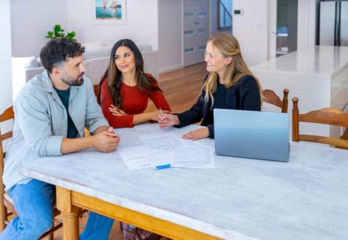 A real estate agent sits at a table with a couple, discussing documents related to a property transaction. The agent smiles while pointing to a document, while the couple listens attentively. This scene highlights a typical negotiation scenario, where clear communication is crucial. Common mistakes in such negotiations could include failing to document agreements or not fully understanding each party's needs.