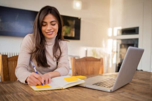 A woman studying at a wooden table with a laptop and notebook reviews example real estate exam questions for her California real estate exam.